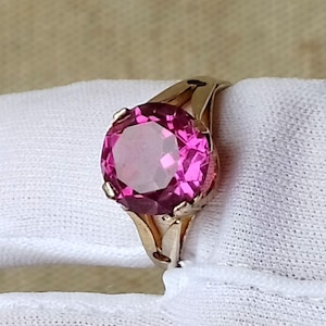 Könnte beinhalten: Ein goldener Ring mit einem großen, runden, facettierten rosa Edelstein. Der Edelstein ist der Mittelpunkt, eingebettet in ein klassisches, elegantes Design. Der Ring hat eine schlichte, aber raffinierte Form.