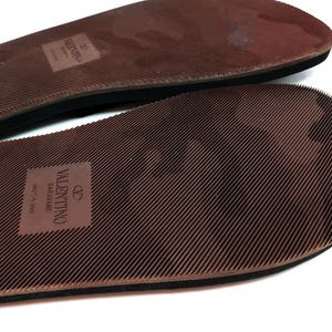 May include: Close-up of the brown rubber soles of a pair of sandals. The soles have a textured pattern and the text "VALENTINO GARAVANI MADE IN ITALY" is printed on the bottom sole.
