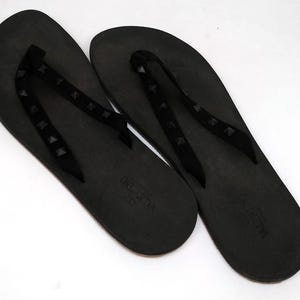 May include: A pair of black flip-flops with studded straps. The straps are made of a black material and have small, black studs. The flip-flops are made of a black rubber material and have the word "VALENTINO" printed on the sole.
