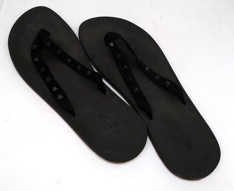 May include: A pair of black flip-flops with studded straps. The straps are made of a black material and have small, black studs. The flip-flops are made of a black rubber material and have the word "VALENTINO" printed on the sole.