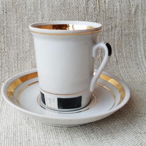 May include: White porcelain teacup and saucer set with gold trim. The cup has a cylindrical shape with a curved handle. The saucer and the rim of the cup are accented with a gold band. A black and white design is on the saucer.