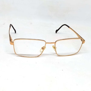 May include: Gold-coloured eyeglasses with rectangular lenses and thin frames. The temples are dark brown and curve down to hook behind the ears. The glasses have a small decorative element on the outer corners of the frames.