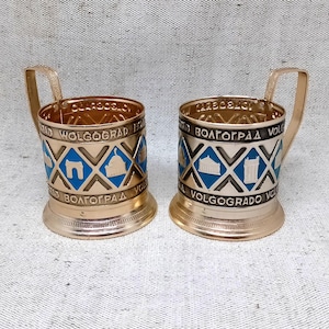 May include: Two gold-colored metal cup holders with handles. Each holder features blue and white geometric designs and the word "VOLGOGRAD" in a circular pattern. The cup holders are likely vintage or souvenir items.