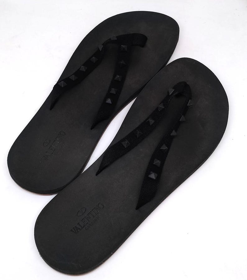 May include: A pair of black leather flip-flops with black studded straps and the Valentino Garavani logo.
