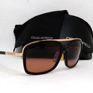 May include: Gold-rimmed sunglasses with brown lenses and a black frame. The sunglasses have the word "DSQUARED2" printed on the frame.