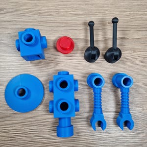 May include: An assortment of blue and black plastic toy parts, including blocks, connectors, and handles, are arranged on a wooden surface. A red circular piece is also present. The parts appear to be 3D-printed and are designed for construction or assembly.