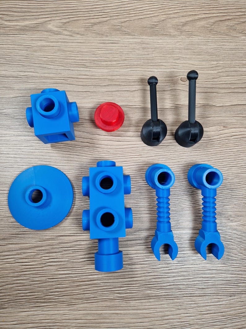 May include: An assortment of blue and black plastic toy parts, including blocks, connectors, and handles, are arranged on a wooden surface. A red circular piece is also present. The parts appear to be 3D-printed and are designed for construction or assembly.
