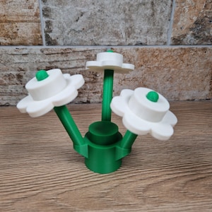 May include: A 3D-printed decorative flower arrangement. The arrangement features three white, daisy-like flowers with green centers and stems. The base is green, and the flowers are arranged in a cluster. The object is made of plastic.