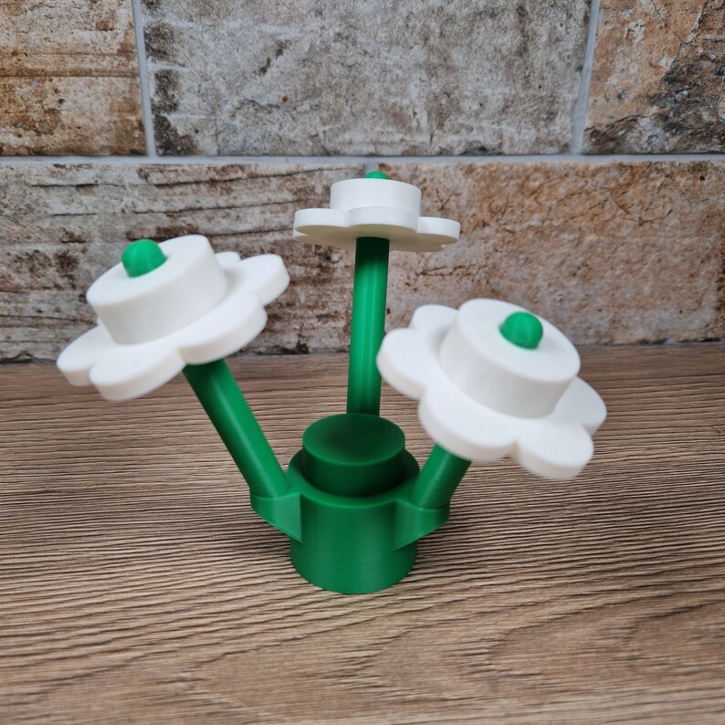 May include: A 3D-printed decorative flower arrangement. The arrangement features three white, daisy-like flowers with green centers and stems. The base is green, and the flowers are arranged in a cluster. The object is made of plastic.