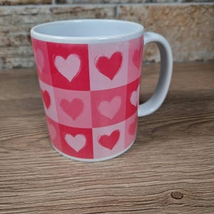 May include: A white ceramic mug with a pink and red heart pattern. The design features a grid of squares, each containing a heart in varying shades of pink. The mug has a white handle and a white interior.