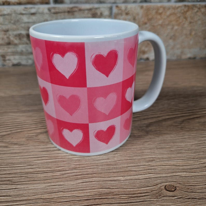 May include: A white ceramic mug with a pink and red heart pattern. The design features a grid of squares, each containing a heart in varying shades of pink. The mug has a white handle and a white interior.