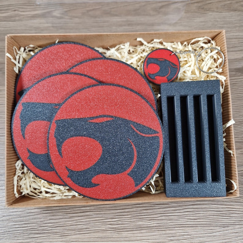 May include: A gift set featuring Thundercats-themed items. Includes four red and black coasters, a keychain, and a small, dark gray rectangular holder. The items are arranged in a brown box with straw-like filler.