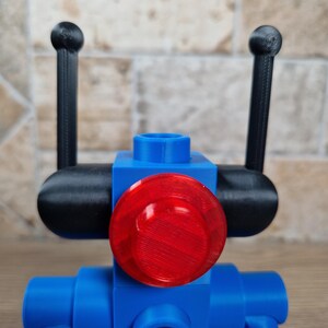 May include: A blue robot toy with a red circular light and black antennae. The robot is constructed from geometric shapes, with a cylindrical body and a round head. The antennae are thin and end in small spheres.