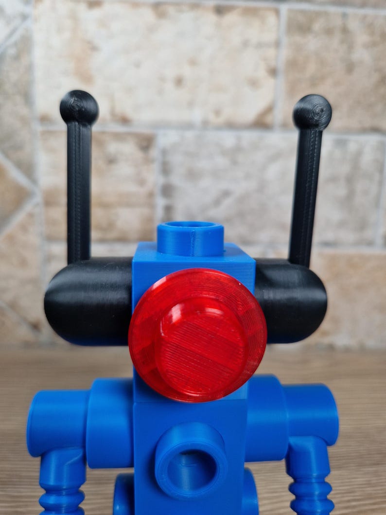 May include: A blue robot toy with a red circular light and black antennae. The robot is constructed from geometric shapes, with a cylindrical body and a round head. The antennae are thin and end in small spheres.