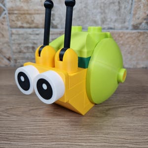 Puede incluir: Un caracol de juguete amarillo y verde hecho de bloques de construcci&oacute;n. El caracol tiene ojos grandes con bordes blancos y pupilas negras, dos antenas negras y una concha redondeada. El juguete est&aacute; dise&ntilde;ado para parecerse a un caracol de dibujos animados.