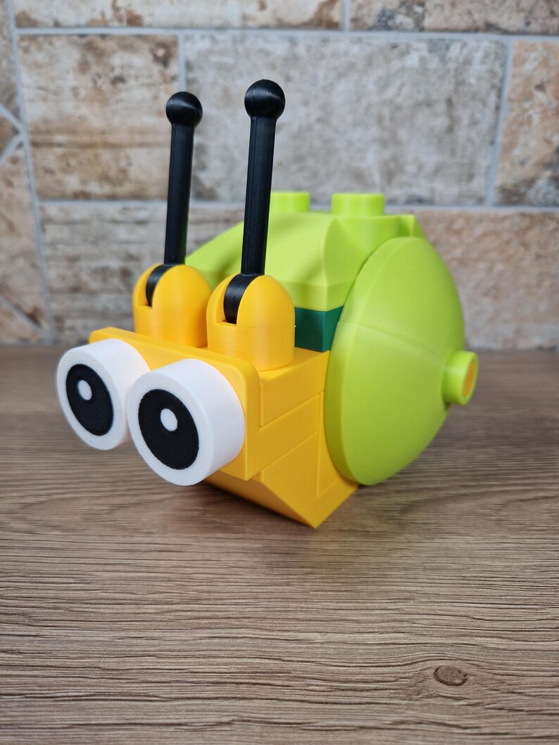 Puede incluir: Un caracol de juguete amarillo y verde hecho de bloques de construcci&oacute;n. El caracol tiene ojos grandes con bordes blancos y pupilas negras, dos antenas negras y una concha redondeada. El juguete est&aacute; dise&ntilde;ado para parecerse a un caracol de dibujos animados.