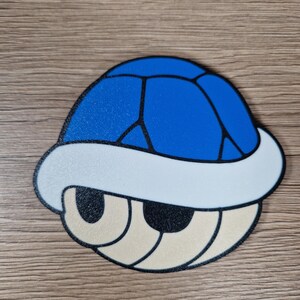 K&ouml;nnte beinhalten: Eine blaue, wei&szlig;e und beige Cartoon-Schildkr&ouml;tenpanzer. Die Schale ist blau mit schwarzen Linien, einem wei&szlig;en Rand und beigen Augen. Das Design ist eine stilisierte Darstellung eines Videospiel-Objekts.