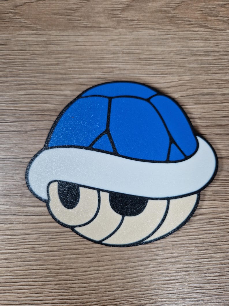 K&ouml;nnte beinhalten: Eine blaue, wei&szlig;e und beige Cartoon-Schildkr&ouml;tenpanzer. Die Schale ist blau mit schwarzen Linien, einem wei&szlig;en Rand und beigen Augen. Das Design ist eine stilisierte Darstellung eines Videospiel-Objekts.