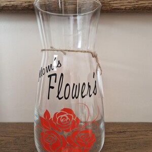 May include: A clear glass vase with a brown twine bow and a black vinyl decal that says "Mom's Flowers" with three red roses.