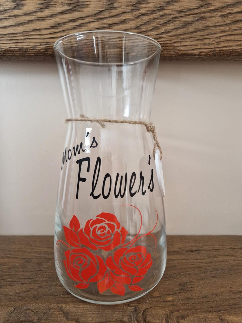 May include: A clear glass vase with a brown twine bow and a black vinyl decal that says "Mom's Flowers" with three red roses.