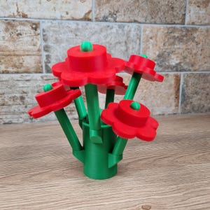 May include: A bouquet of red, flower-shaped objects with green stems and small green accents. The flowers are arranged in a green vase-like base. The arrangement is set on a wooden surface with a brick-like background.