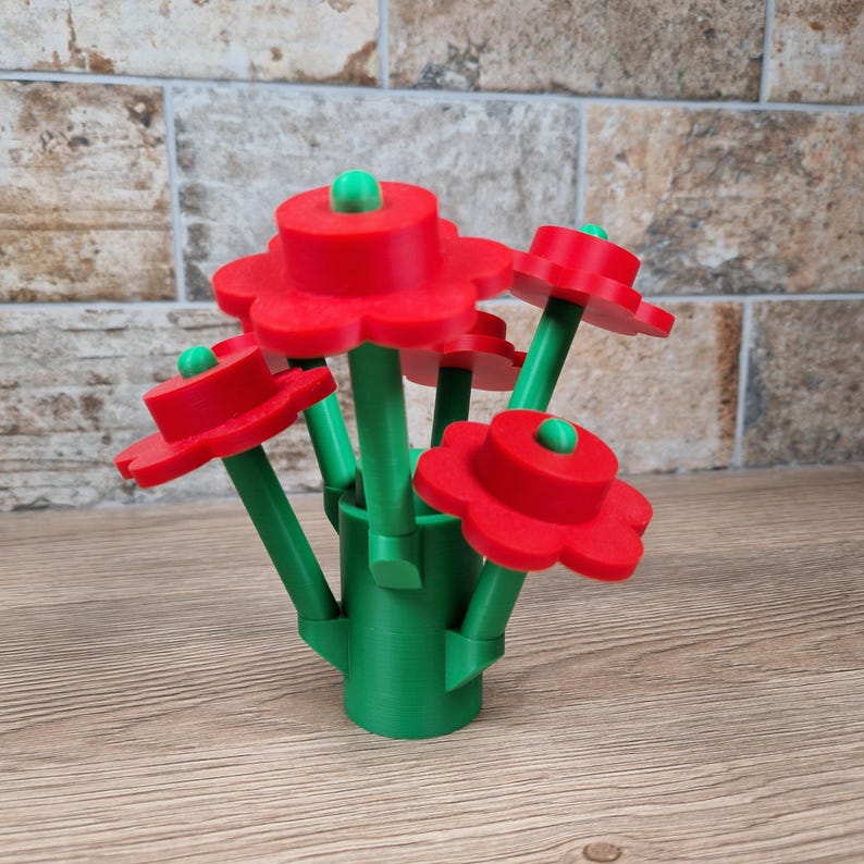May include: A bouquet of red, flower-shaped objects with green stems and small green accents. The flowers are arranged in a green vase-like base. The arrangement is set on a wooden surface with a brick-like background.