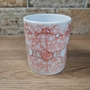 May include: A white ceramic mug with a pink heart and bubble design. The mug features a heart rate monitor graphic with a large heart in the center. Small hearts are strung across the top of the mug.