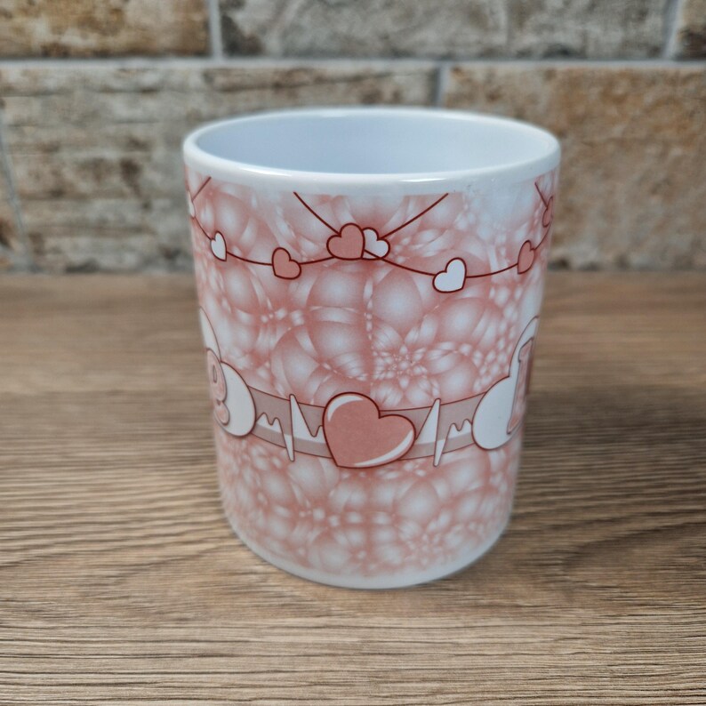 May include: A white ceramic mug with a pink heart and bubble design. The mug features a heart rate monitor graphic with a large heart in the center. Small hearts are strung across the top of the mug.