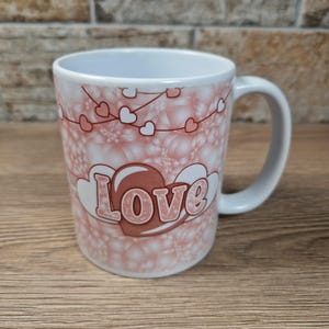 May include: White ceramic mug with a pink and white heart design and the word "Love" in a decorative font. The mug has a white handle and is set against a brick wall and wooden surface.
