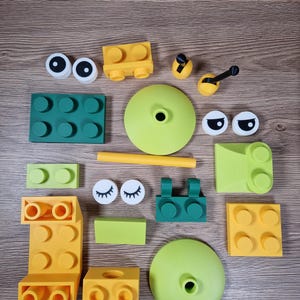 Puede incluir: Una colecci&oacute;n de bloques de construcci&oacute;n y accesorios coloridos en varias formas y tama&ntilde;os. El conjunto incluye piezas amarillas, verdes y blancas, como componentes en forma de ojo, bases redondas y bloques rectangulares, dispuestos sobre una superficie de madera.