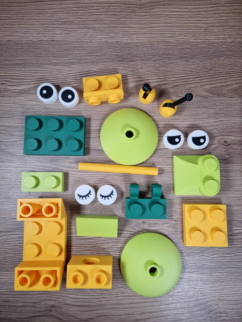 Puede incluir: Una colecci&oacute;n de bloques de construcci&oacute;n y accesorios coloridos en varias formas y tama&ntilde;os. El conjunto incluye piezas amarillas, verdes y blancas, como componentes en forma de ojo, bases redondas y bloques rectangulares, dispuestos sobre una superficie de madera.