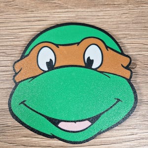 Puede incluir: Una cara de dibujos animados verde y marr&oacute;n con una boca sonriente, ojos blancos y una m&aacute;scara marr&oacute;n. La cara tiene un contorno negro y probablemente sea un personaje de una serie animada popular.