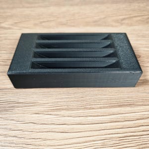 May include: A black, rectangular object with a series of angled slots. The object is made of a textured material and sits on a light brown wooden surface. The slots are evenly spaced and create a geometric pattern.