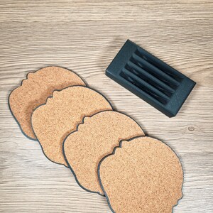 Puede incluir: Juego de cuatro posavasos de corcho con forma de cabeza, con borde azul oscuro, y un soporte gris oscuro. Los posavasos son de color marr&oacute;n natural. El soporte est&aacute; dise&ntilde;ado para guardarlos.