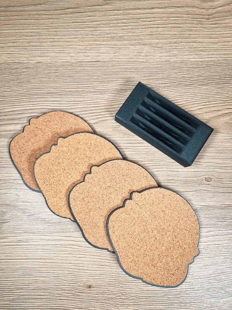 Puede incluir: Juego de cuatro posavasos de corcho con forma de cabeza, con borde azul oscuro, y un soporte gris oscuro. Los posavasos son de color marr&oacute;n natural. El soporte est&aacute; dise&ntilde;ado para guardarlos.