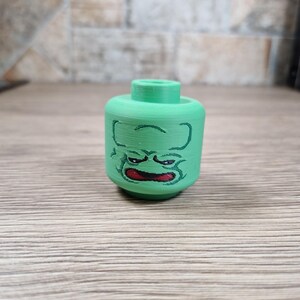 May include: A green, 3D-printed toy head with a cartoonish, angry face. The face features black outlines and a red mouth. The head is cylindrical with a raised, circular top. The toy is designed to be compatible with building block sets.