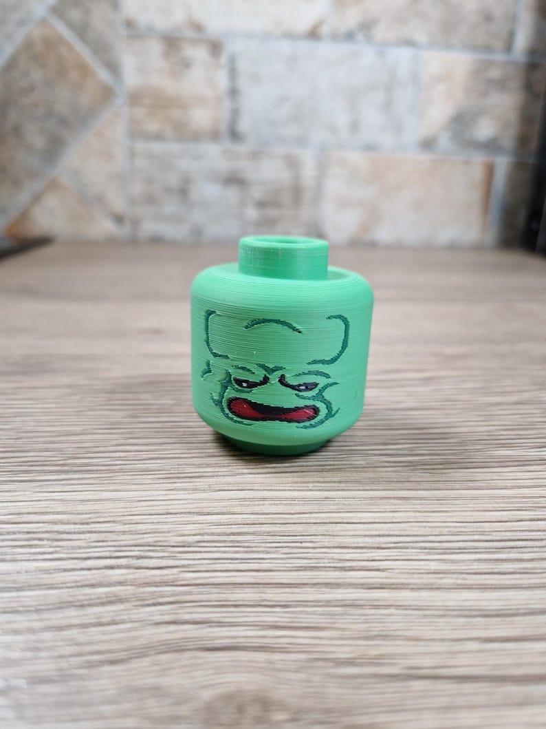 May include: A green, 3D-printed toy head with a cartoonish, angry face. The face features black outlines and a red mouth. The head is cylindrical with a raised, circular top. The toy is designed to be compatible with building block sets.
