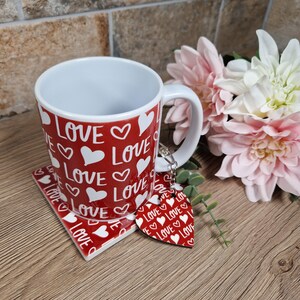 K&ouml;nnte beinhalten: Ein rotes Keramik-Set mit Tasse, Untersetzer und herzf&ouml;rmigem Schl&uuml;sselanh&auml;nger. Tasse und Untersetzer sind mit dem Wort "LOVE" und wei&szlig;en Herzen verziert. Der Schl&uuml;sselanh&auml;nger hat das gleiche Design. Ein Strau&szlig; rosa und wei&szlig;er Blumen liegt daneben.