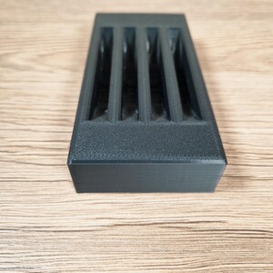 May include: A rectangular, dark gray 3D-printed object with four vertical slots. The object has a textured surface and is set against a light brown wooden background. The design suggests a storage or organizational product.