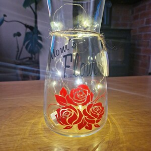 May include: A clear glass vase with a string of lights inside. The vase has a red rose design and the text "Mom's Flowers" on the front.
