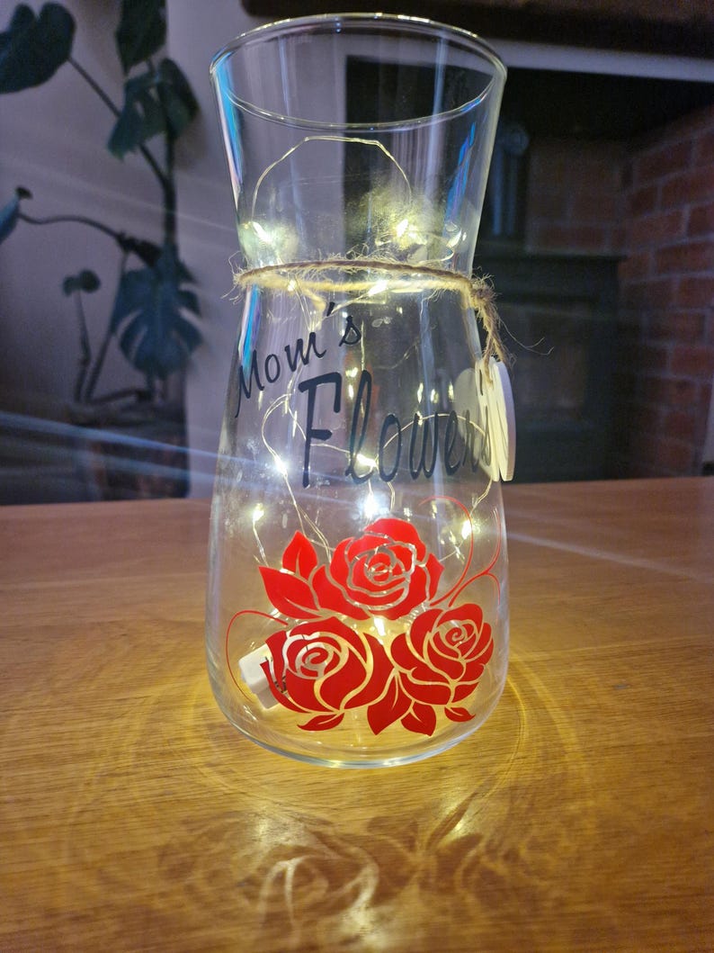 May include: A clear glass vase with a string of lights inside. The vase has a red rose design and the text "Mom's Flowers" on the front.