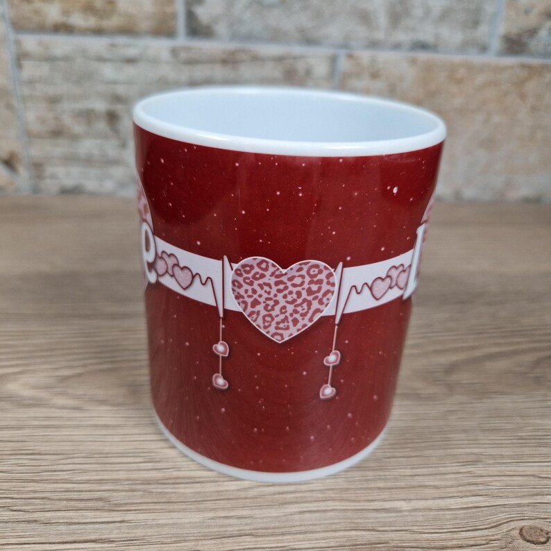 May include: A red ceramic mug with a white interior. The mug features a white band with the word "Mom" and a leopard print heart design. The mug has a speckled pattern and is suitable for hot or cold beverages.