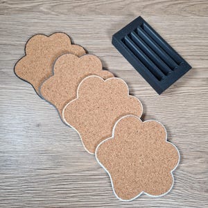 Op de afbeelding: Een set pootvormige kurken onderzetters met witte rand, geplaatst op een houten oppervlak. Een kleine, zwarte houder met vier sleuven staat ernaast. De onderzetters zijn lichtbruin van kleur.