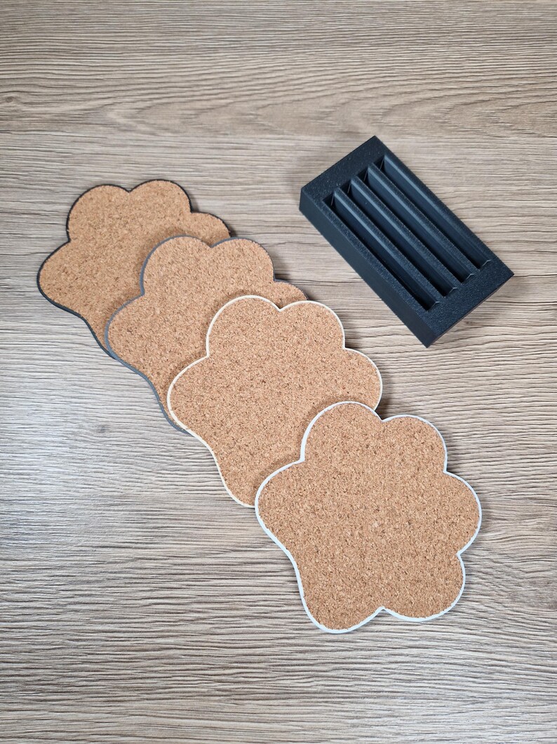 Op de afbeelding: Een set pootvormige kurken onderzetters met witte rand, geplaatst op een houten oppervlak. Een kleine, zwarte houder met vier sleuven staat ernaast. De onderzetters zijn lichtbruin van kleur.