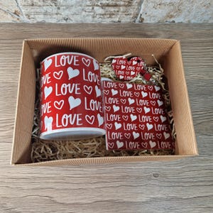 K&ouml;nnte beinhalten: Ein Geschenkset in einem Karton, mit einer roten Tasse und einem passenden Artikel, beide mit dem Wort "LOVE" und wei&szlig;en Herzen verziert. Die Artikel sind in dekorativem Stroh gebettet.