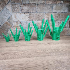 May include: A set of five green 3D-printed cactus-shaped holders. Each holder has a cylindrical base with multiple upward-pointing, rod-like structures. The holders vary in size, with the smallest on the left and the largest on the right.