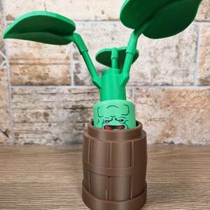 May include: A green, plant-like sculpture with large leaves and a face emerging from a brown barrel-shaped base. The leaves are large and rounded, and the face has a surprised expression. The sculpture is made of plastic.