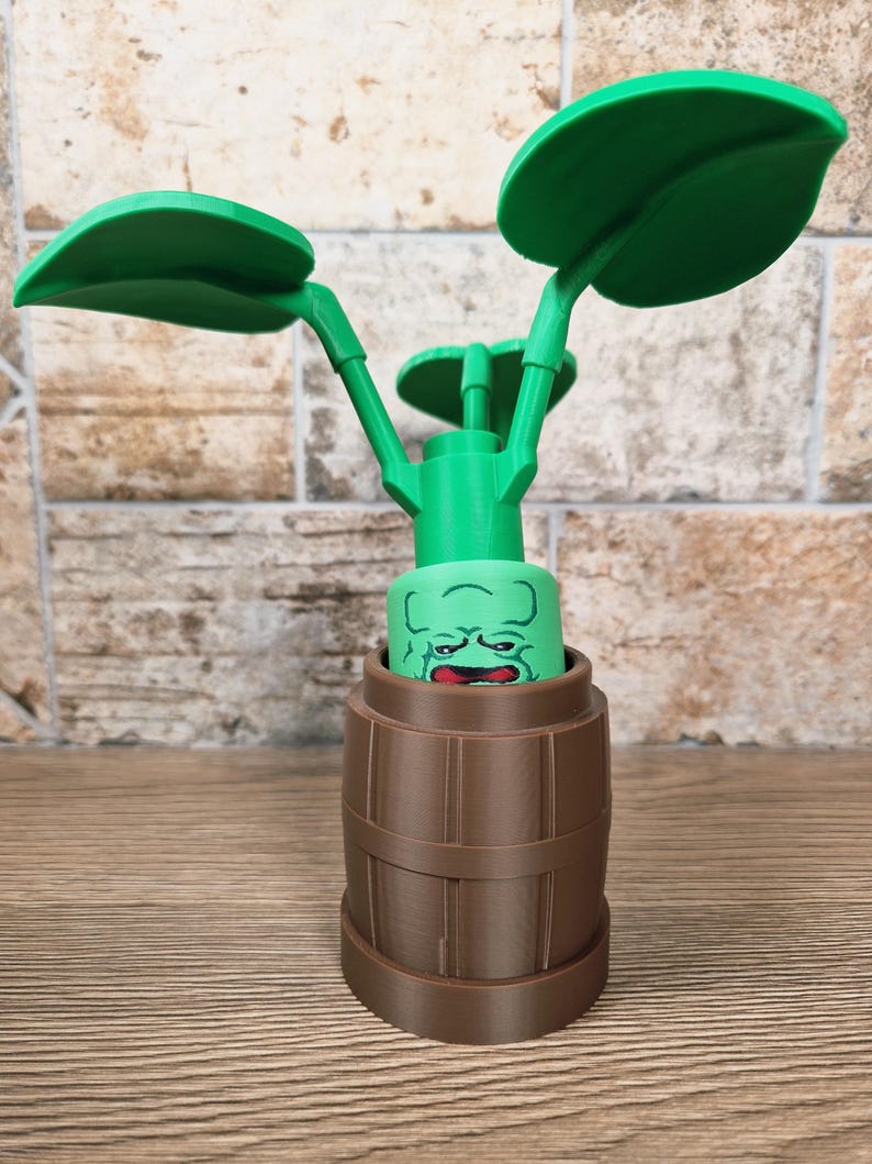 May include: A green, plant-like sculpture with large leaves and a face emerging from a brown barrel-shaped base. The leaves are large and rounded, and the face has a surprised expression. The sculpture is made of plastic.