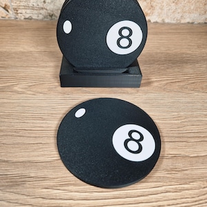 May include: Black round coasters with a white number 8 and a white dot, resembling a billiard ball. The coasters are stacked on a black square holder. The coasters are on a wooden surface.