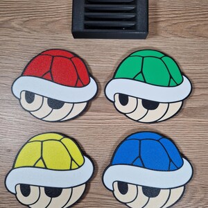 K&ouml;nnte beinhalten: Vier bunte Schildkr&ouml;tenpanzer-Untersetzer in Rot, Gr&uuml;n, Gelb und Blau, mit wei&szlig;em Rand und beigem Gesichtsdesign. Ein schwarzer rechteckiger Halter befindet sich &uuml;ber den Untersetzern. Diese Untersetzer sind eine lustige Erg&auml;nzung f&uuml;r jedes Zuhause.